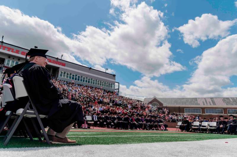 a man in a graduation gown and cap sitting in a chair in front of a crowd of people