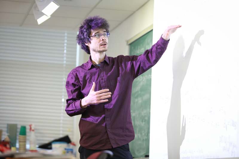 a man with purple hair and purple shirt pointing at a chalkboard