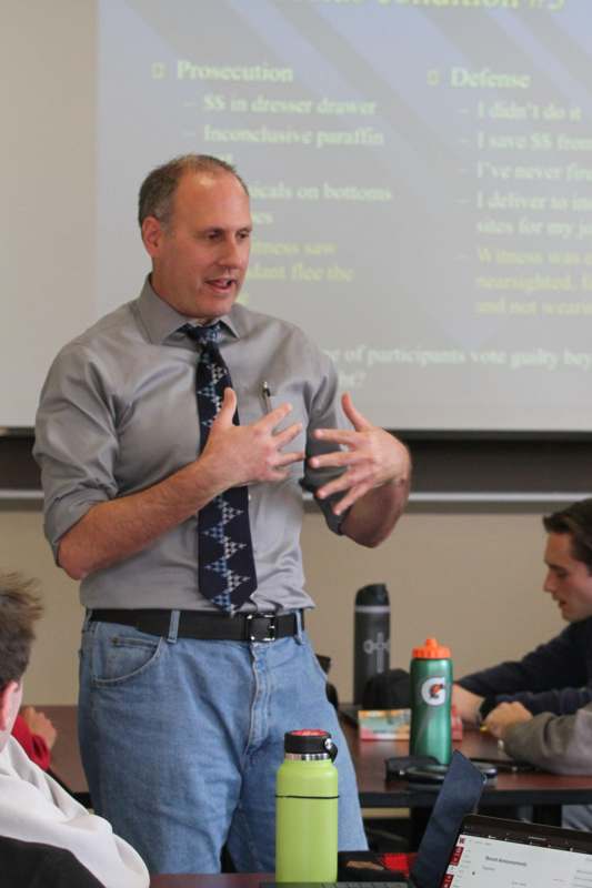 a man in a tie and jeans speaking in front of a screen