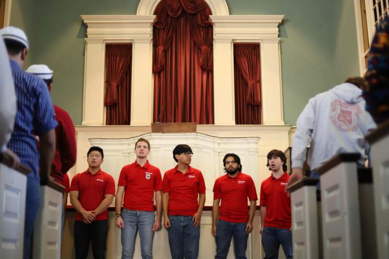 a group of men in red shirts standing in front of a man standing in front of a stage