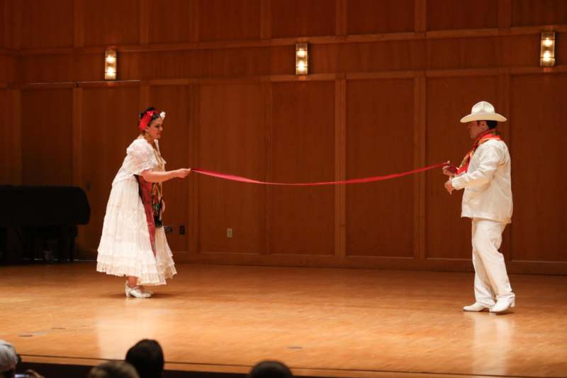 a man and woman in white dress and hat on stage with a red ribbon