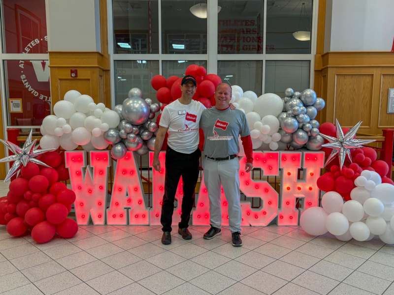 two men standing in front of a large sign with balloons