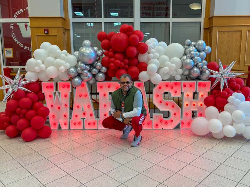 a man squatting in front of a large sign with red and white balloons