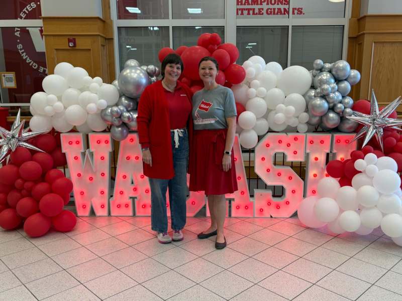 two women standing in front of a large sign with red and white balloons
