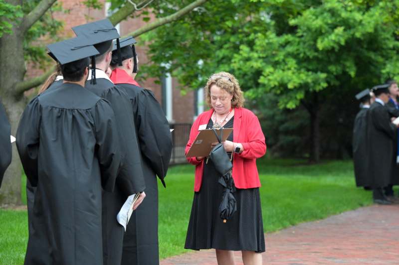 a woman in a red jacket holding a folder with a group of people in caps and gowns