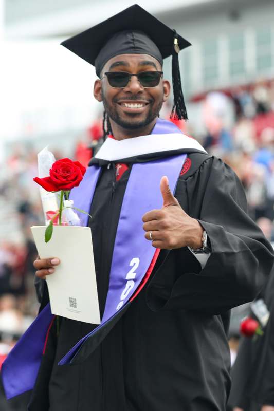 a man in a graduation gown holding a rose and giving a thumbs up
