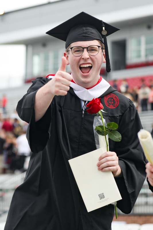 a man in a graduation gown holding a rose and giving a thumbs up