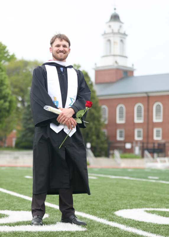 a man in a graduation gown holding a rose and standing on a football field