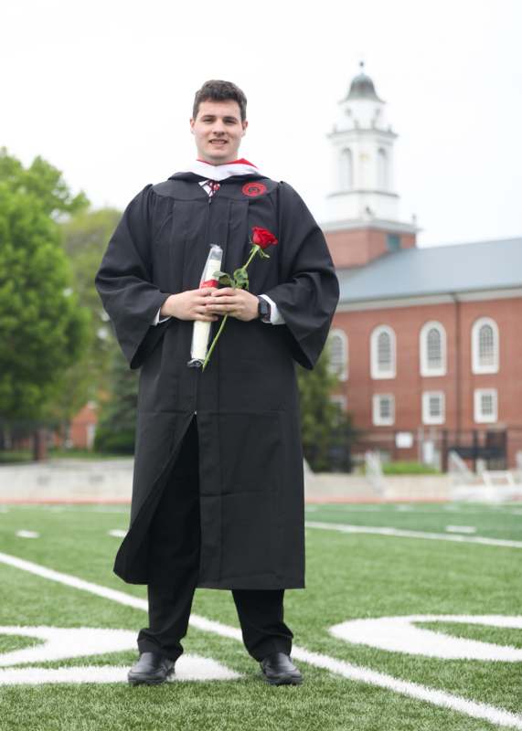 a man in a graduation gown holding a rose and standing on a football field