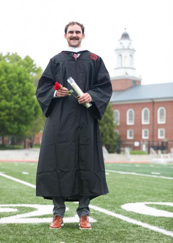 a man in a graduation gown holding a rose and standing on a football field